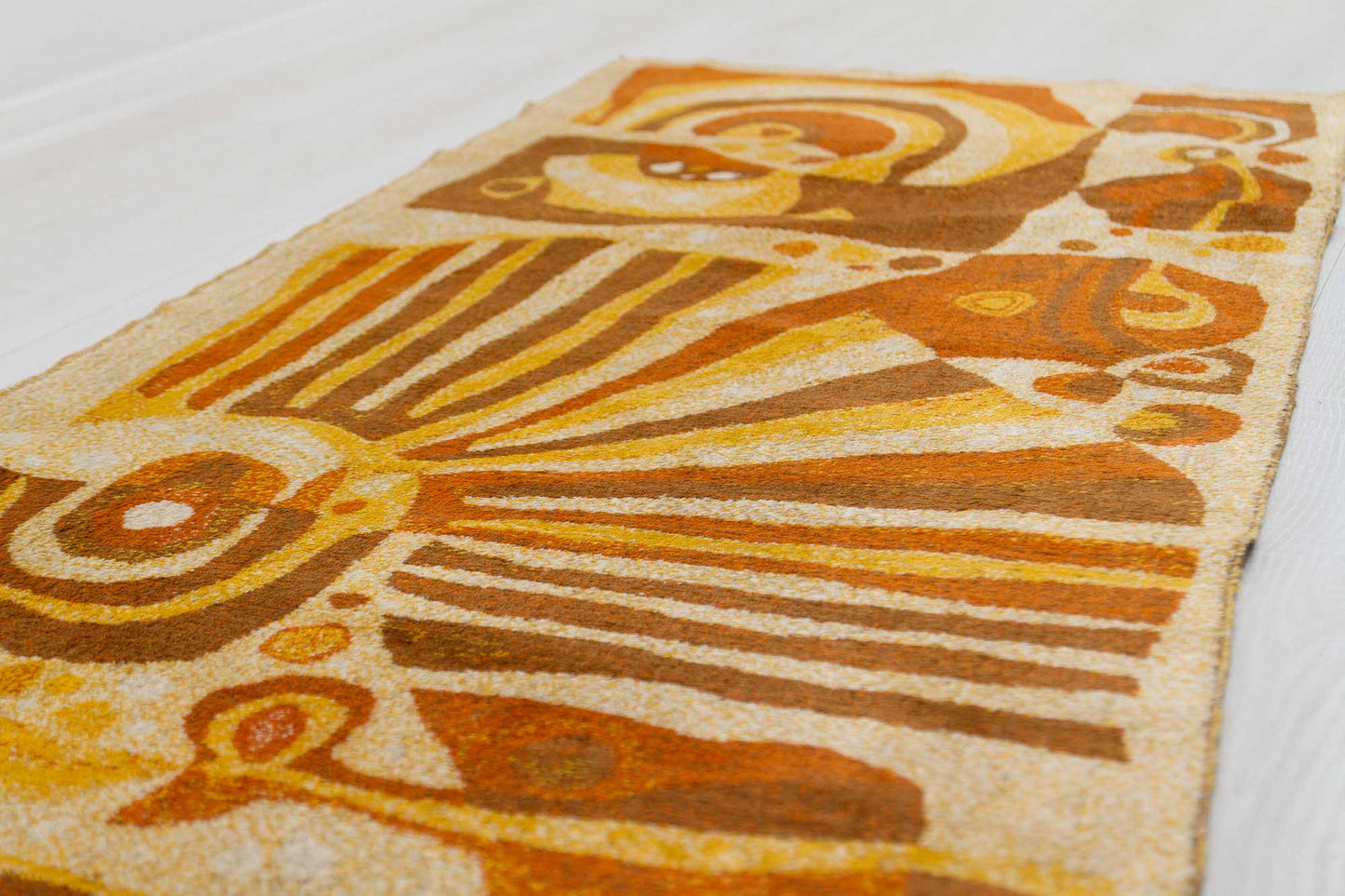 angled view of a 1960s vintage mid century modern wall tapestry laid on a white floor, showcasing the organic abstract design in a typically mid-century color scheme of orange, brown and beige