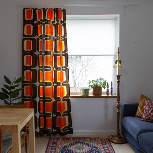 Mid century modern curtain panel on white rod next to a window, glowing 70s orange and chocolate brown retro print.