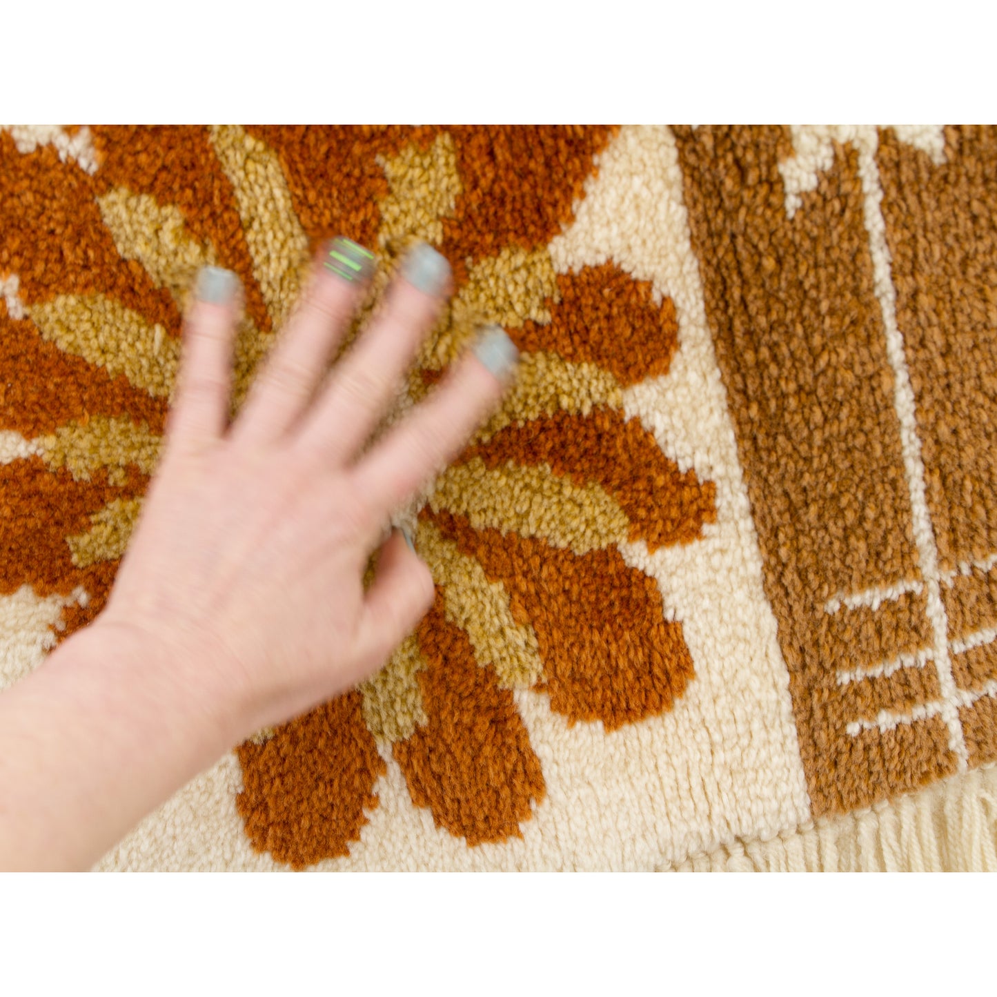 Hand hovering over the soft surface of the tapestry. Color detail showing the gradient of 1970s earth tones: Cream background with floral motifs in burnt orange, spicy mustard, and deep brown.