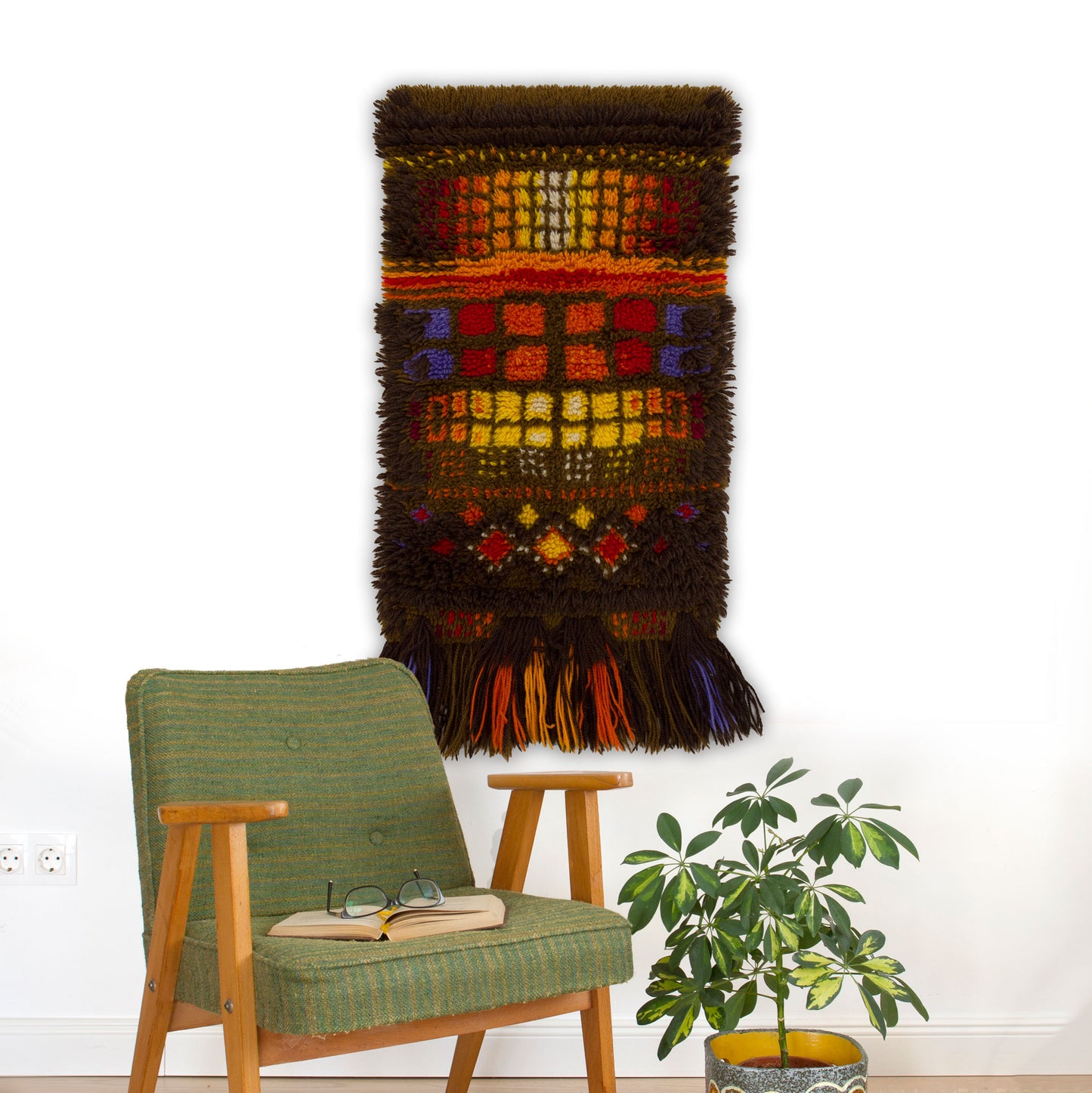 A colorful 1970s Smyrnafix latch hook tapestry hanging on a white wall above a green mid-century armchair, featuring rich dark brown, bright orange, and yellow pixel-like geometry.