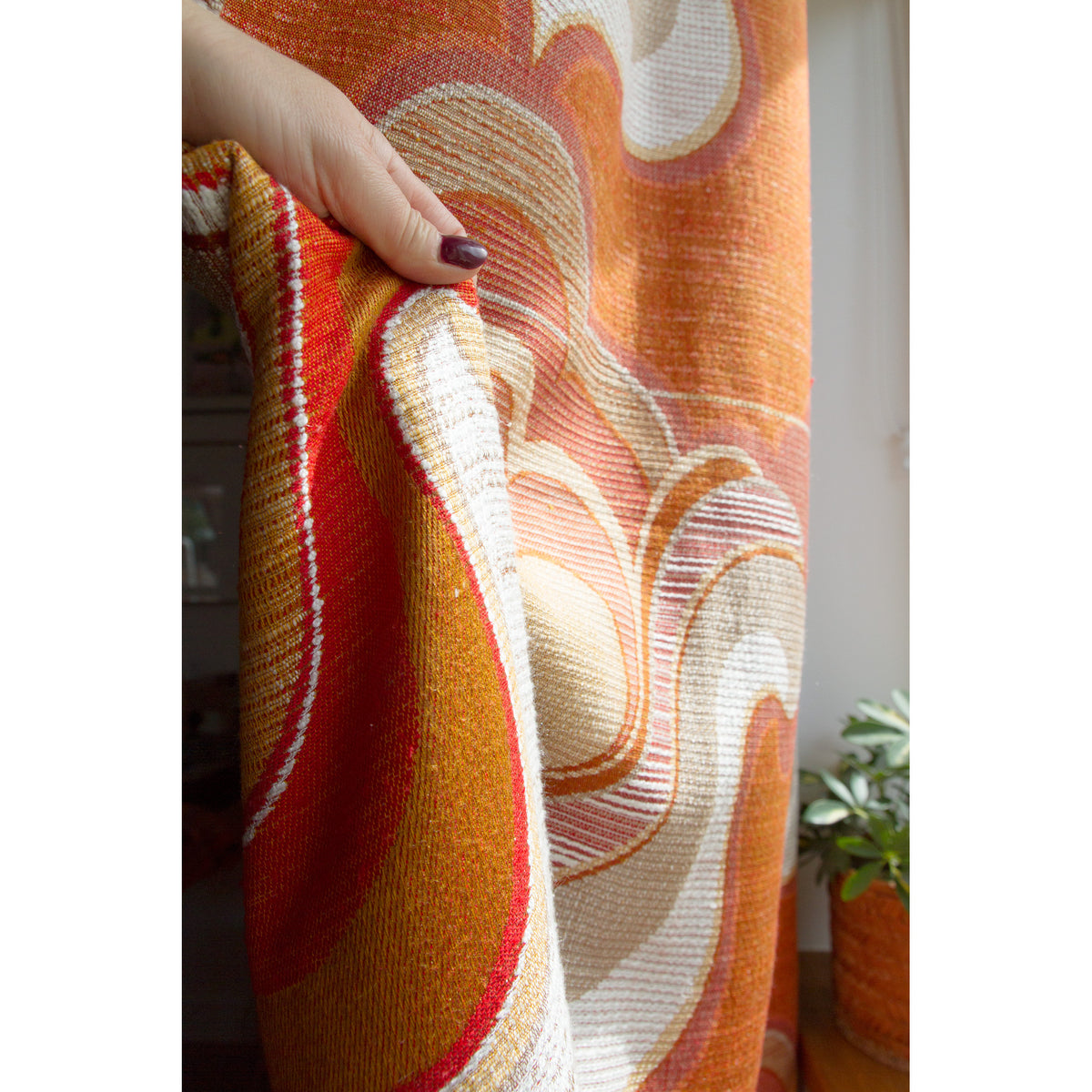 70s curtain panel pulled aside by hand, showing the heavy drape and retro orange red pattern