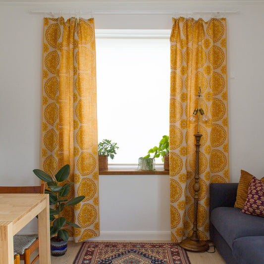 Retro yellow woven curtains framing a window in a boho living room with blue sofa, brass floor lamp and vintage rug.