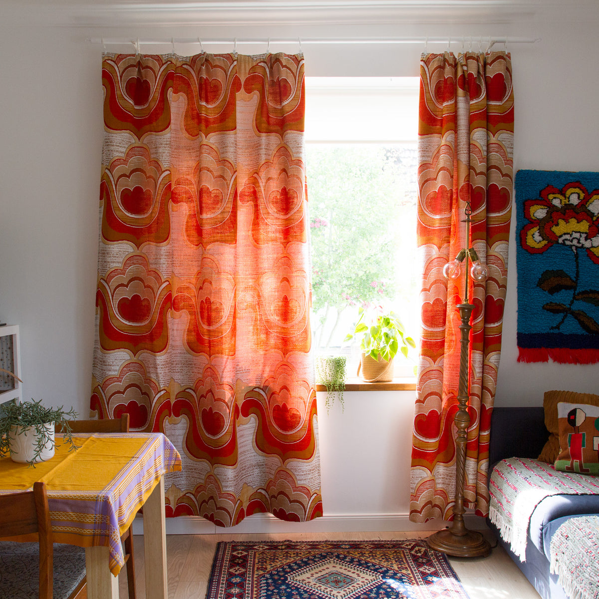 Pair of 1970s vintage curtains with woven orange, red and cream flower power pattern hanging at a window in a retro style living room