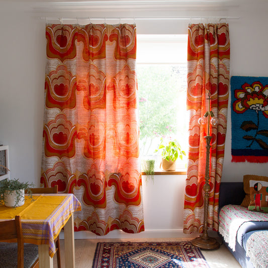 Pair of 1970s vintage curtains with woven orange, red and cream flower power pattern hanging at a window in a retro style living room
