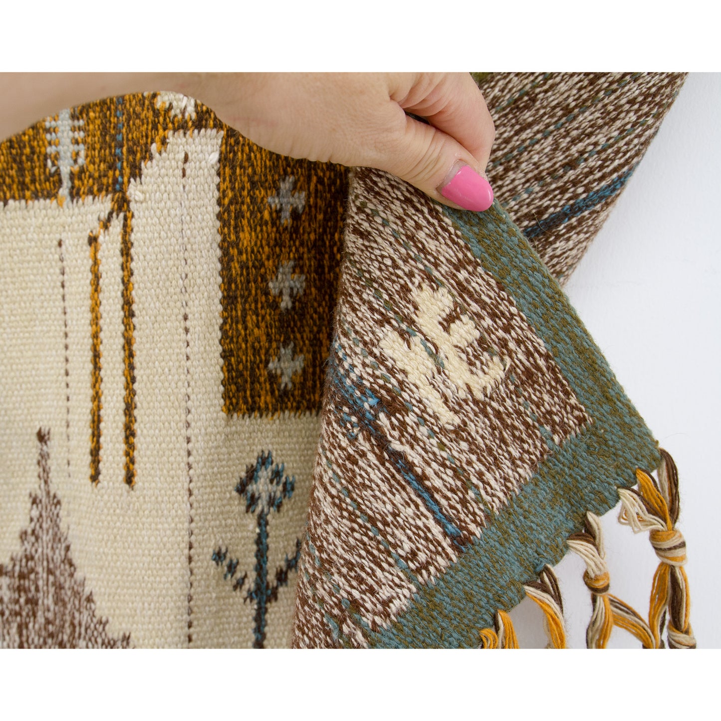 Hand holding the bottom corner of the vintage tapestry, verifying authenticity by highlighting the original artist's signature letters 'NE' proudly woven into the wool fabric.