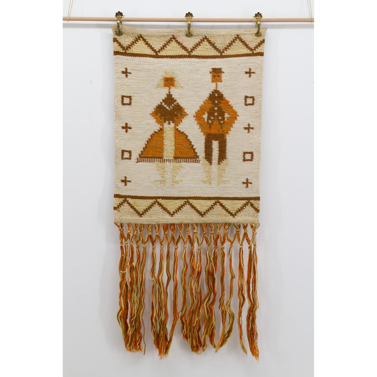 Flat view of a mid-century geometric bohemian tapestry mounted via brass clips on a wooden dowel, featuring natural wool and naive 1970s art motifs.