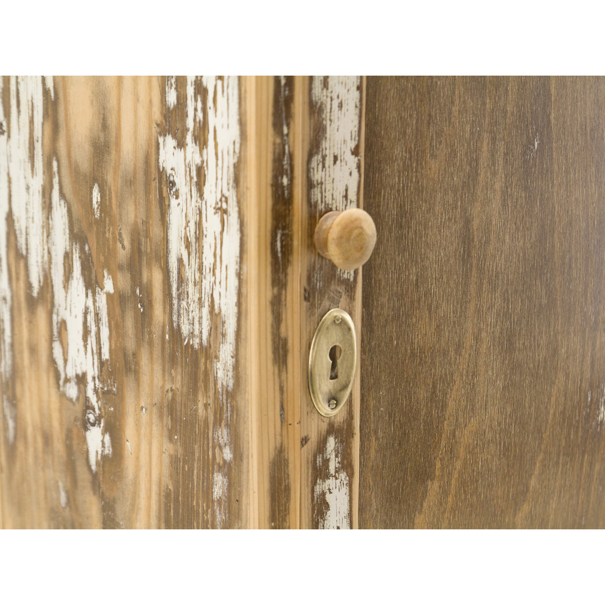 Close-up of a rustic wooden cabinet door with distressed white paint peeling away to reveal natural wood grain, featuring a round wooden knob and an oval brass keyhole. The vintage shaker style furniture is completely refurbished and refinished with premium wood wax for a satin finish.