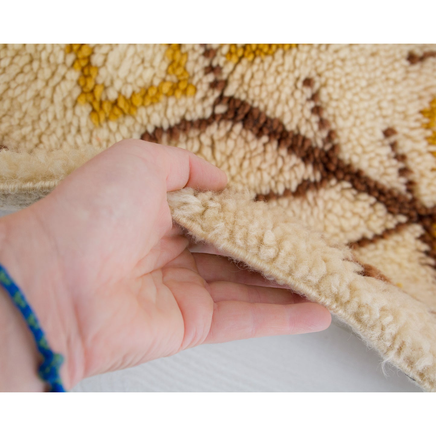 Detailed hand interaction on the carpet edge, proving to vintage buyers the impeccably high quality, densely-packed yarn strands making up the off-white, yellow, and brown pattern.
