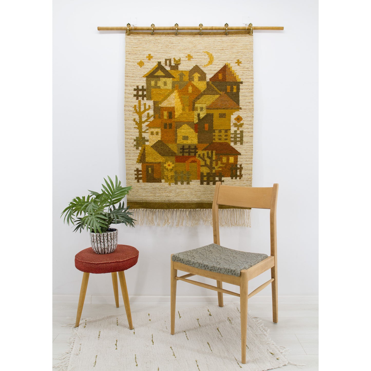 A charming 1970s vintage geometric village tapestry with earth tones hanging elegantly on a bright wall behind a mid-century side chair and green plant.