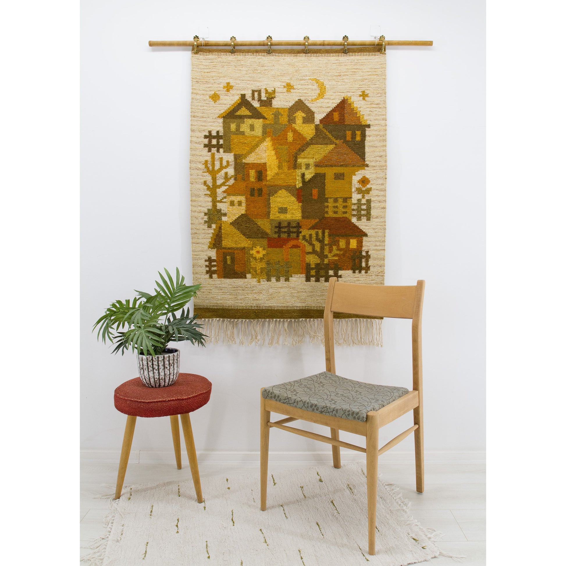 A charming 1970s vintage geometric village tapestry with earth tones hanging elegantly on a bright wall behind a mid-century side chair and green plant.
