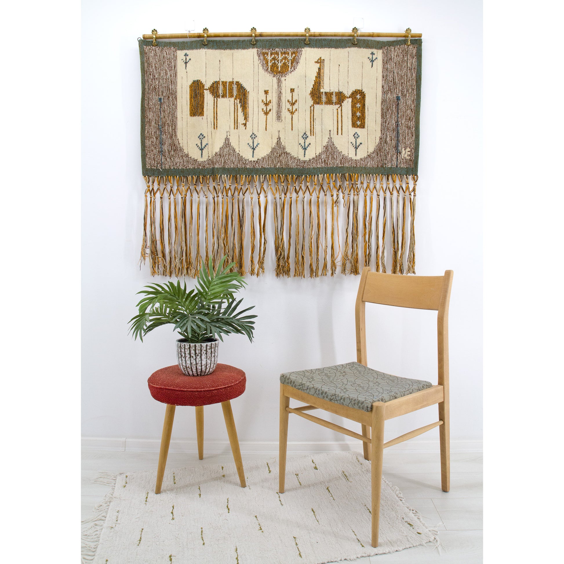 A vintage 1970s Eva Nemeth handwoven wool tapestry with long fringes hanging on a white wall above a mid-century wooden dining chair and green houseplant.