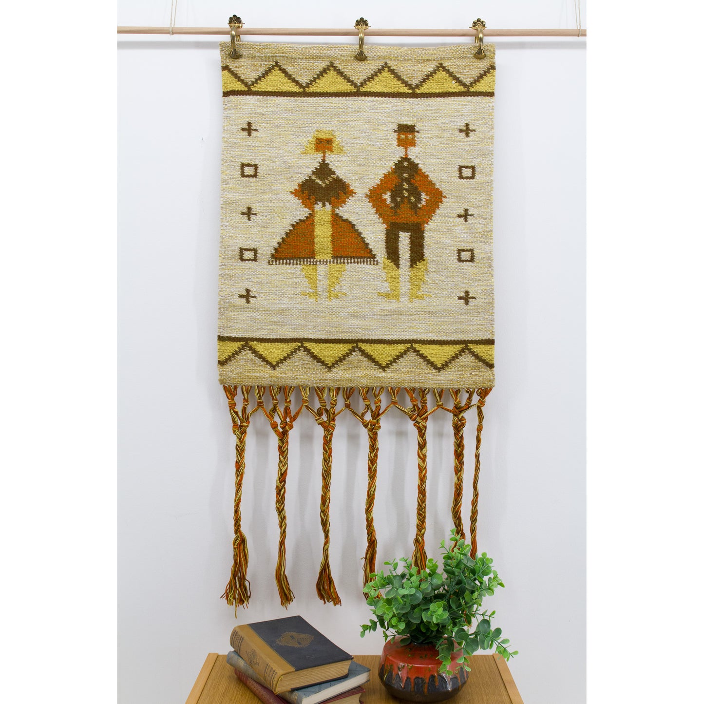 Straight on, brightly lit photo of a vintage mid century folk art wall hanging showing a highly stylized geometric man and woman motif in mustard and earthy terracotta orange.