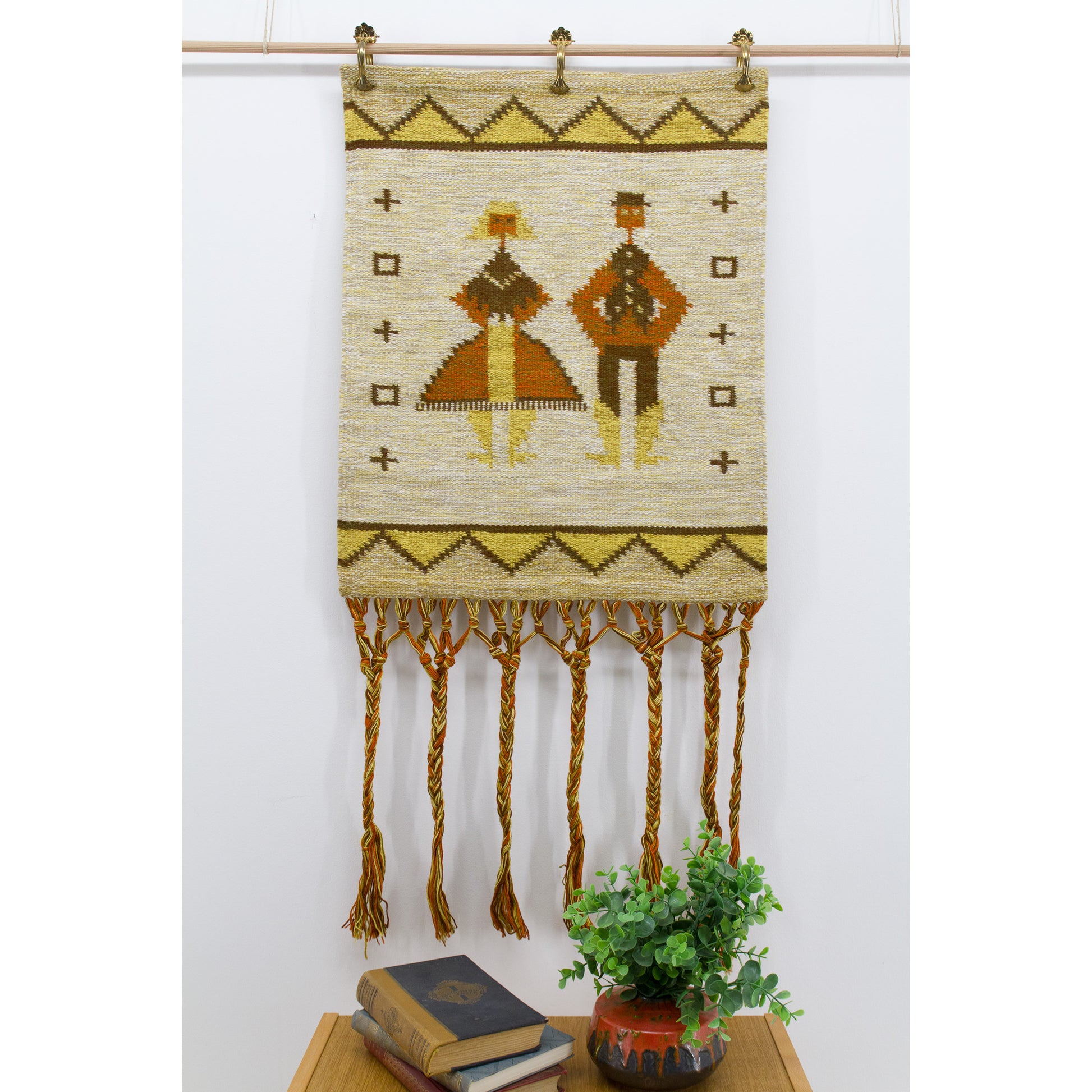 Straight on, brightly lit photo of a vintage mid century folk art wall hanging showing a highly stylized geometric man and woman motif in mustard and earthy terracotta orange.