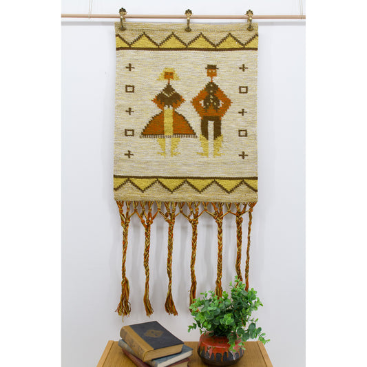 Straight on, brightly lit photo of a vintage mid century folk art wall hanging showing a highly stylized geometric man and woman motif in mustard and earthy terracotta orange.