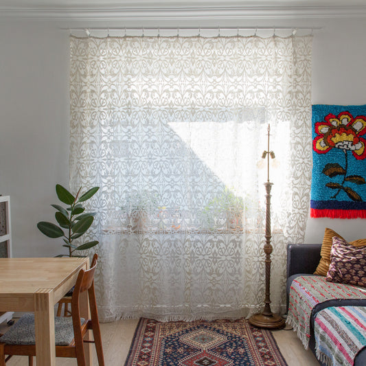 Extra large vintage ivory cotton lace curtain covering a sunny window in a cozy living room with floor lamp and tapestry