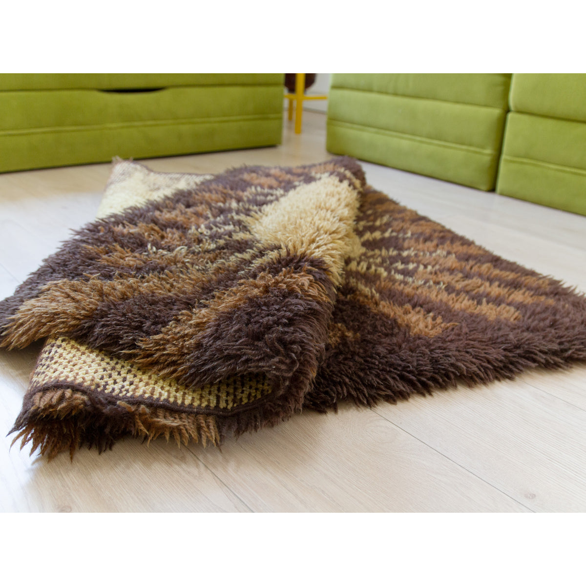 A vintage rya rug with a striped pattern in dark brown, medium brown, and beige, folded on a light wood floor in a mid-century style living room setting with green modular sofas in the background.