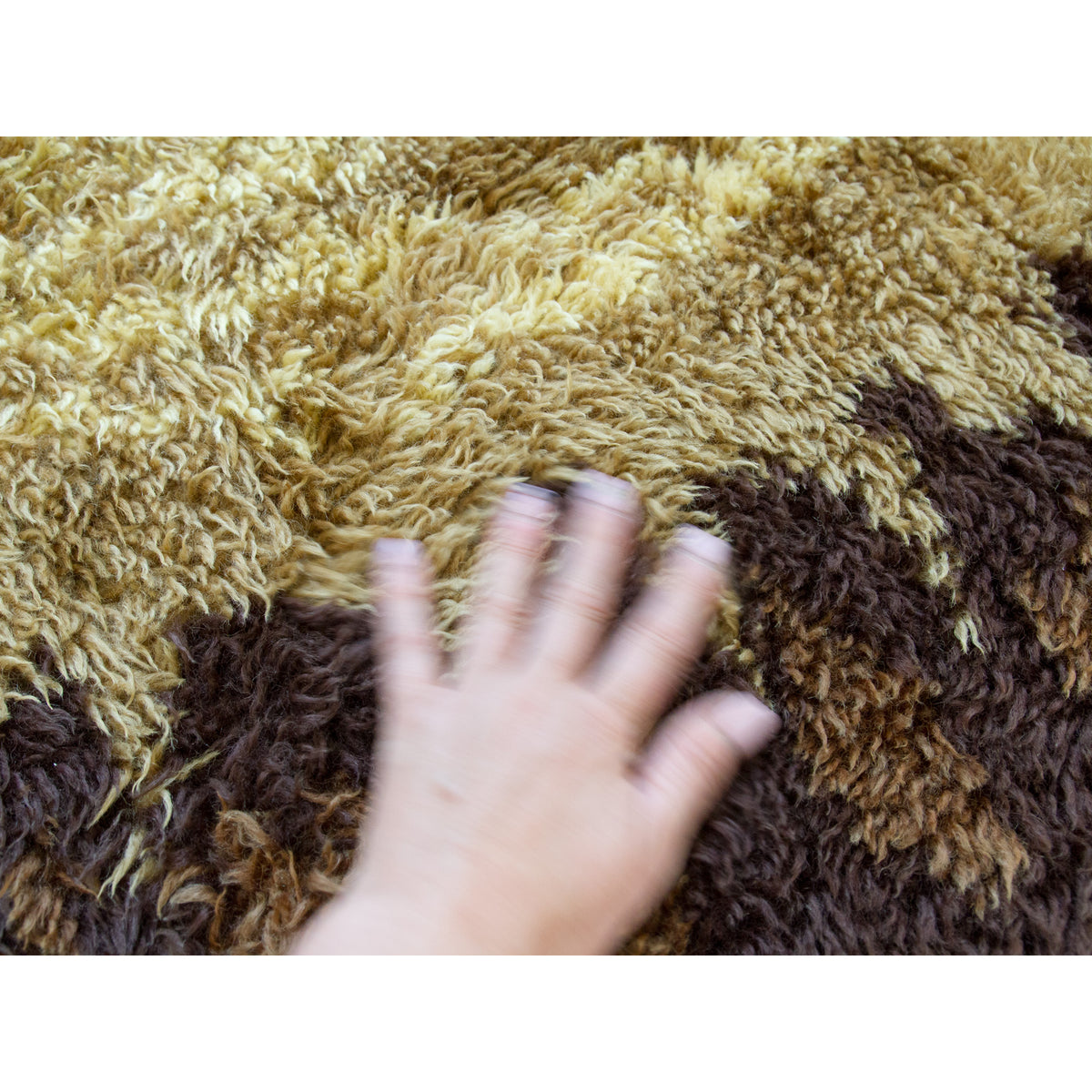 A close-up of a shaggy, long-pile rug in alternating bands of tan/beige and dark brown, with a human hand placed on top to show texture and scale of the vintage rug.