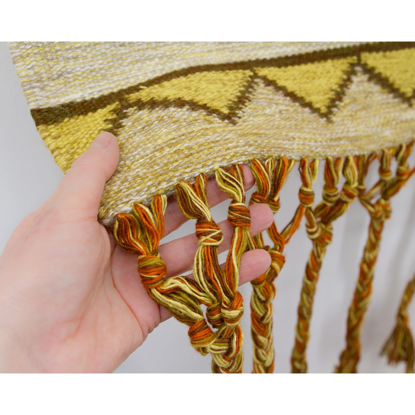 A close-up showing a hand holding the bottom hem of the 1970s wool wall hanging, drawing focus to the high-quality attachment of the long, beautifully braided macrame fringes and warm woven texture.