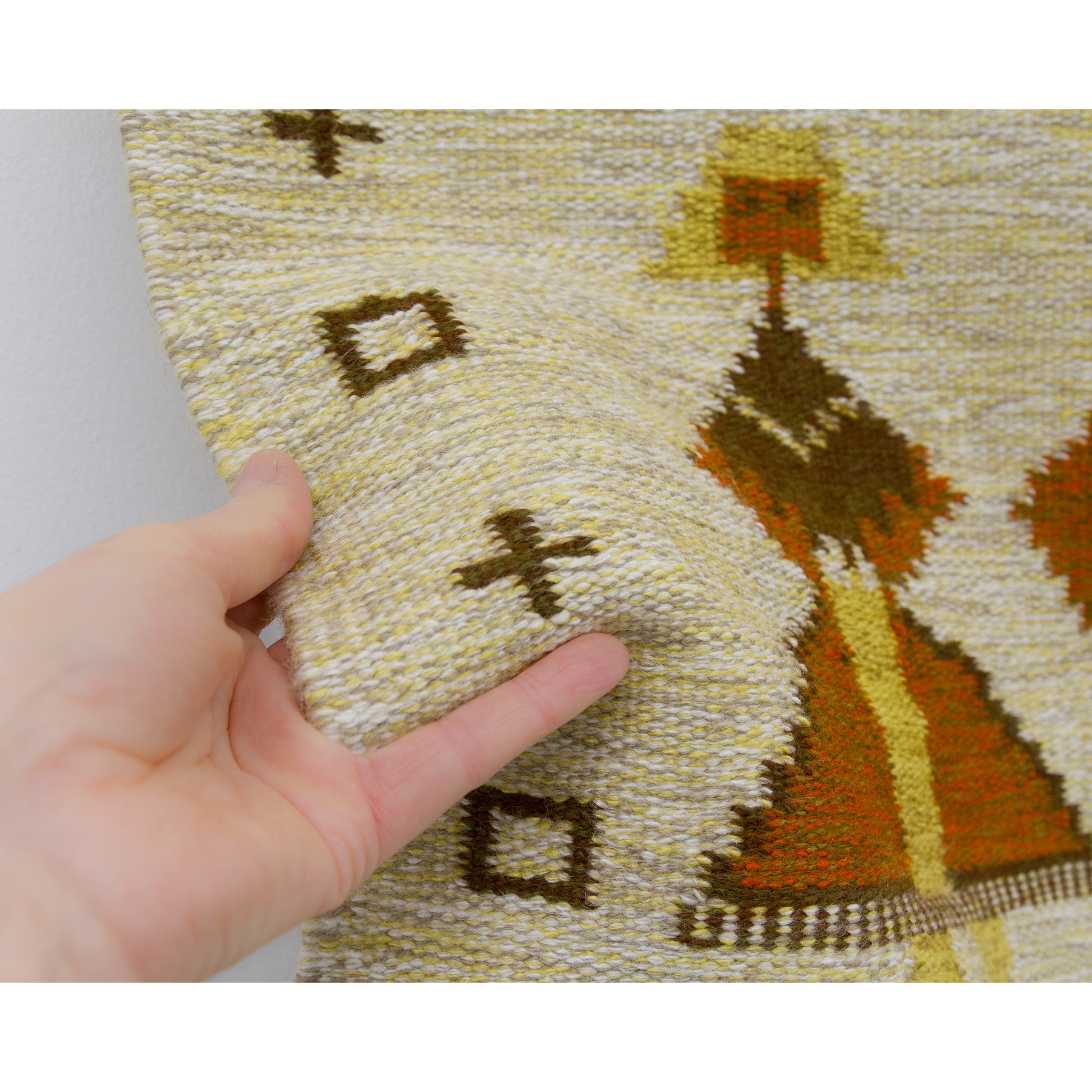 A hand gently lifting the soft woven wool fabric of a 1960s folk art tapestry, showing the high-quality weaving texture and creamy beige cotton warp background.