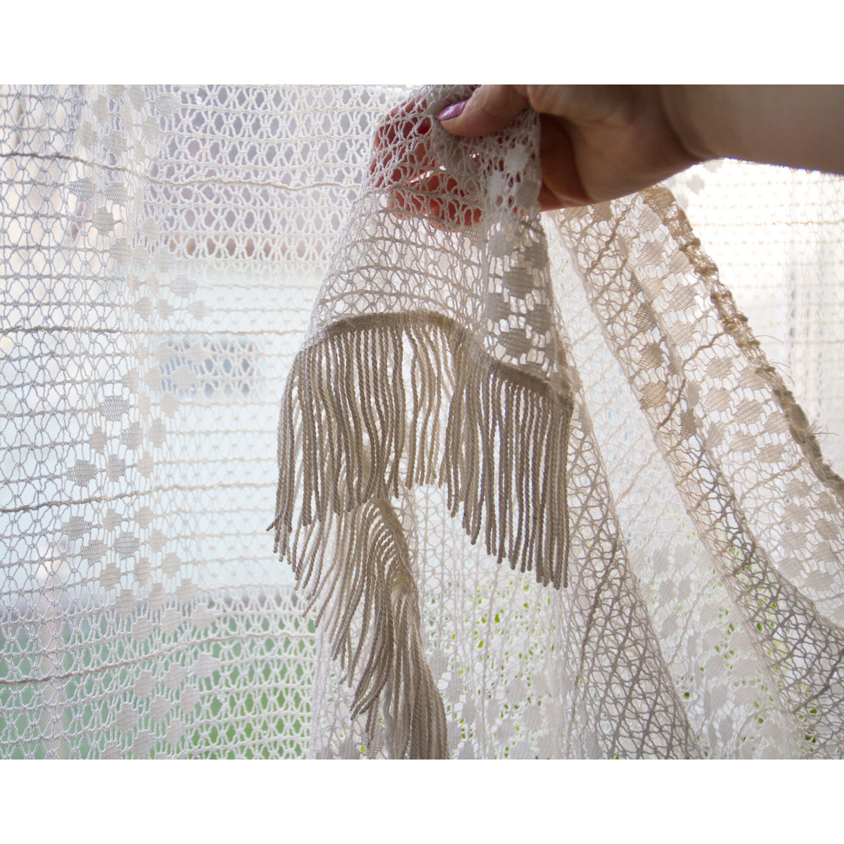 Corner of vintage lace curtain held by hand, showing the long cotton fringe detail