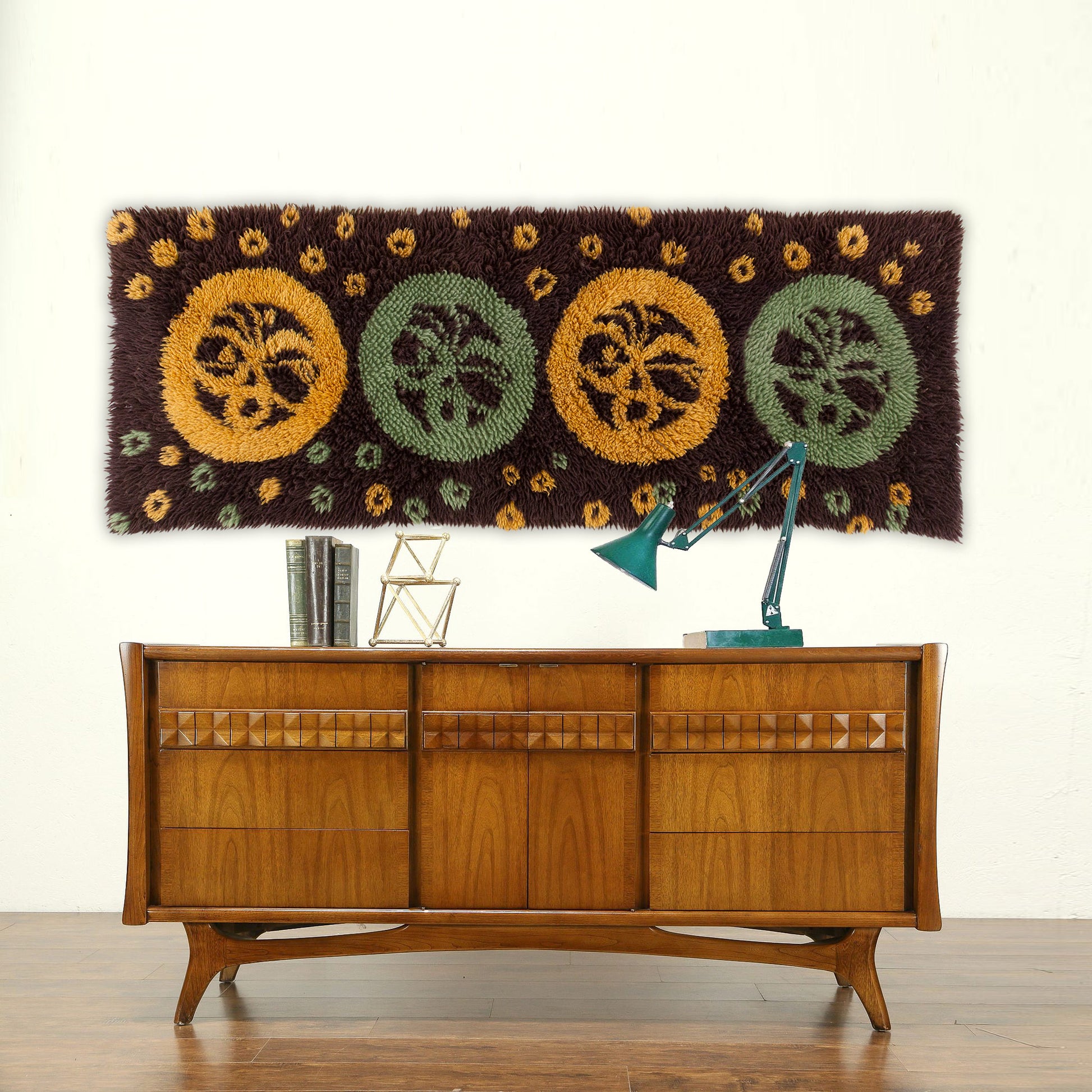 A massive groovy 1970s vintage latch hook tapestry featuring brown, mint green, and mustard circles hung elegantly over a beautiful wooden mid century teak credenza.
