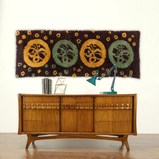 A massive groovy 1970s vintage latch hook tapestry featuring brown, mint green, and mustard circles hung elegantly over a beautiful wooden mid century teak credenza.