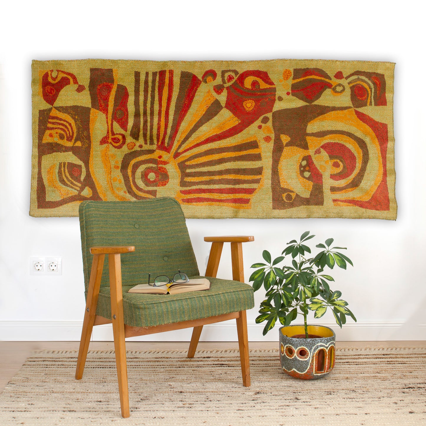 The vintage tapestry styled as a statement piece above a green upholstered armchair. Shows how the warm brown and orange tones complement biophilic, organic mid century modern interiors.