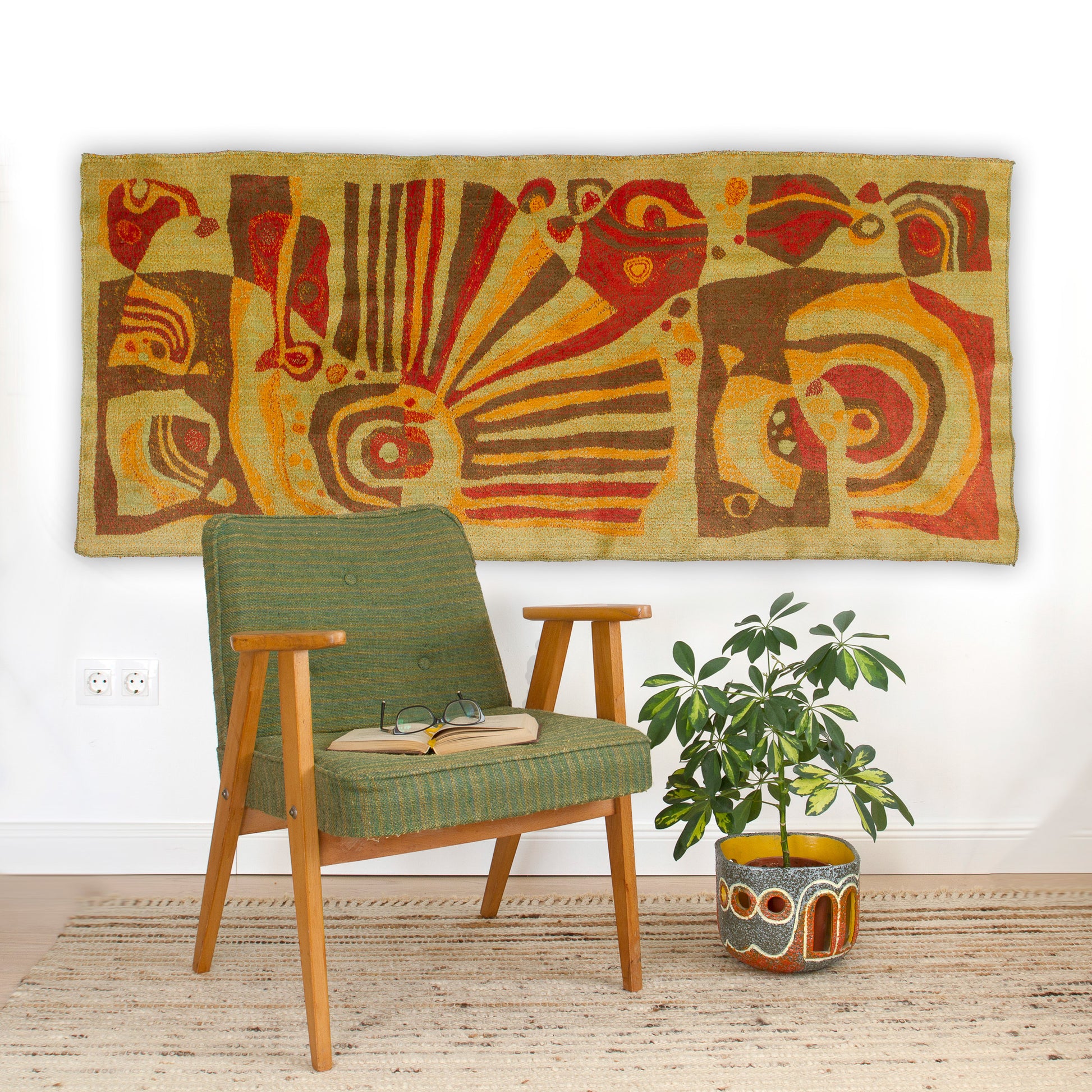 The vintage tapestry styled as a statement piece above a green upholstered armchair. Shows how the warm brown and orange tones complement biophilic, organic mid century modern interiors.