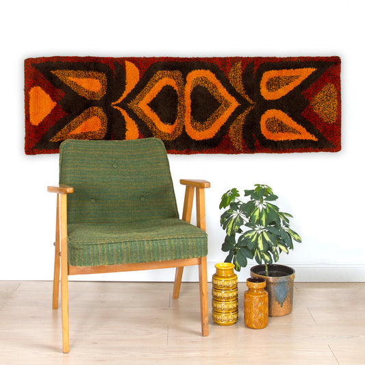 A horizontal 68-inch mid century modern geometric tapestry hung majestically over a green retro armchair, bringing warm acoustic dampening to a room.