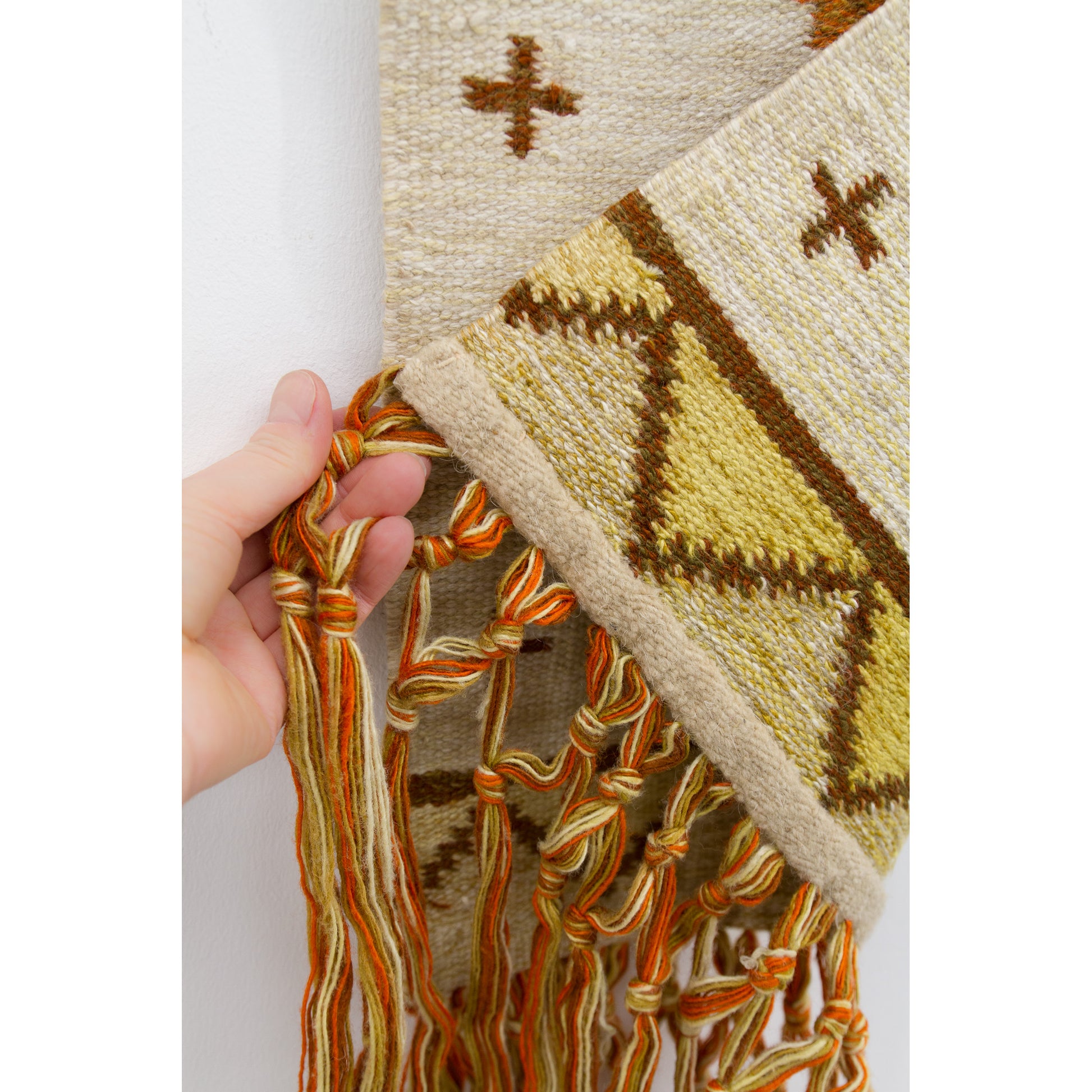 Hand holding the neatly hemmed bottom edge of the woven artwork folded upward, highlighting how securely the knotted macrame fringe is attached.