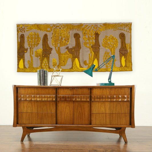 A 67-inch vintage mid century modern tapestry styled flawlessly over a warm wooden teak credenza. Features vibrant mustard yellow and brown organic folk art silhouettes.