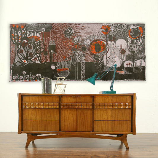 Large vintage 1960s tapestry by László Pécsi styled above a wooden mid-century credenza. The abstract brutalist wall art features a striking, dark moody color palette with vivid orange and white organic shapes.