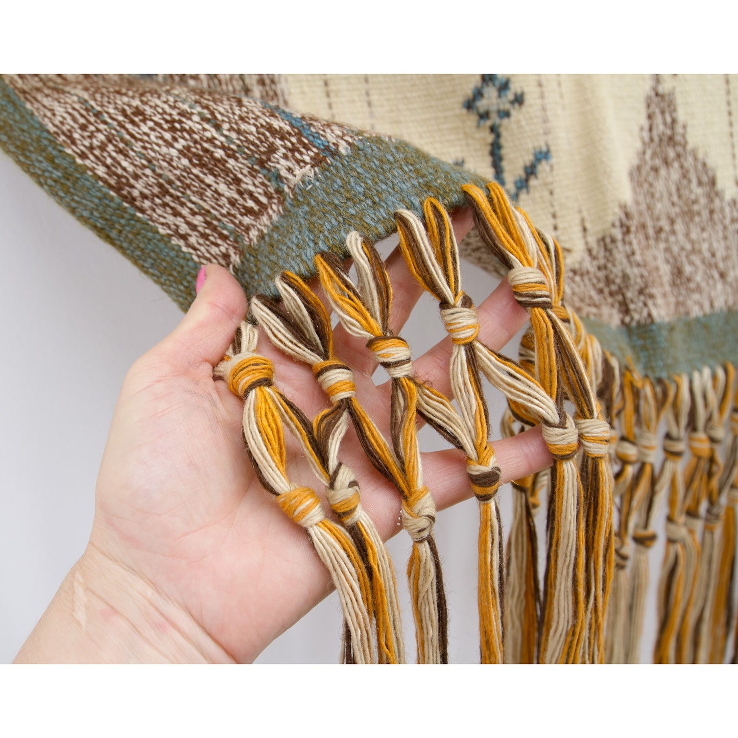 Hand holding the beautifully long macrame fringes, showcasing the wonderfully natural, earthy yarn color palette of creamy beige, brown, and golden orange.