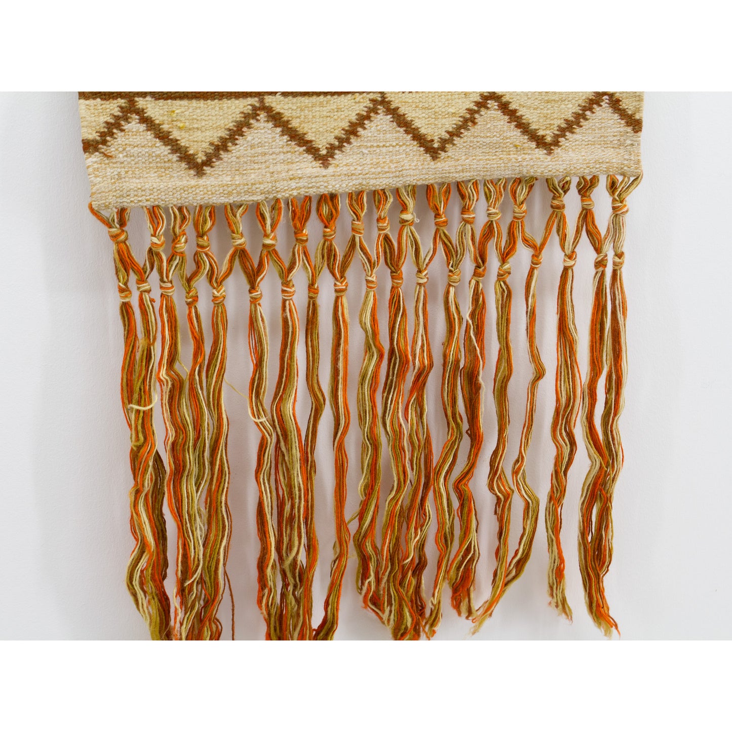 Close up on the long, earthy orange, brown, and beige wool macrame fringes, beautifully handknotted to finish the bottom edge of the textile.