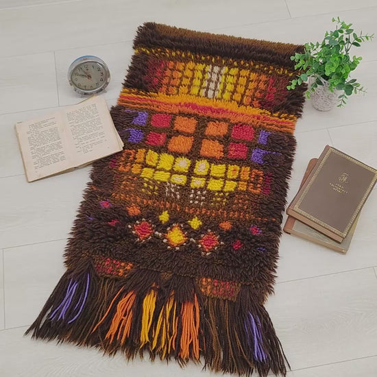 A sweeping video panning over a vintage 70s orange and brown geometric Smyrnafix long pile rug, with hands manipulating the dense, colorful shag fibers to show thickness and lush texture.