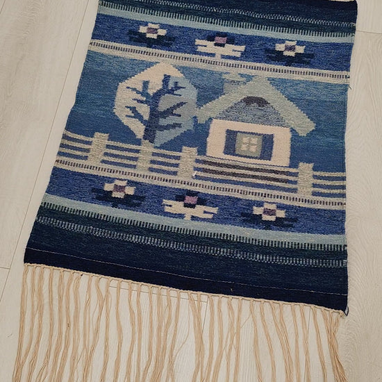 Video showcasing the vintage handwoven wool tapestry laid flat. Closing up on the detailed geometric design of the small house and the tree motives while demonstrating the soft wool texture of the high quality flatweave.