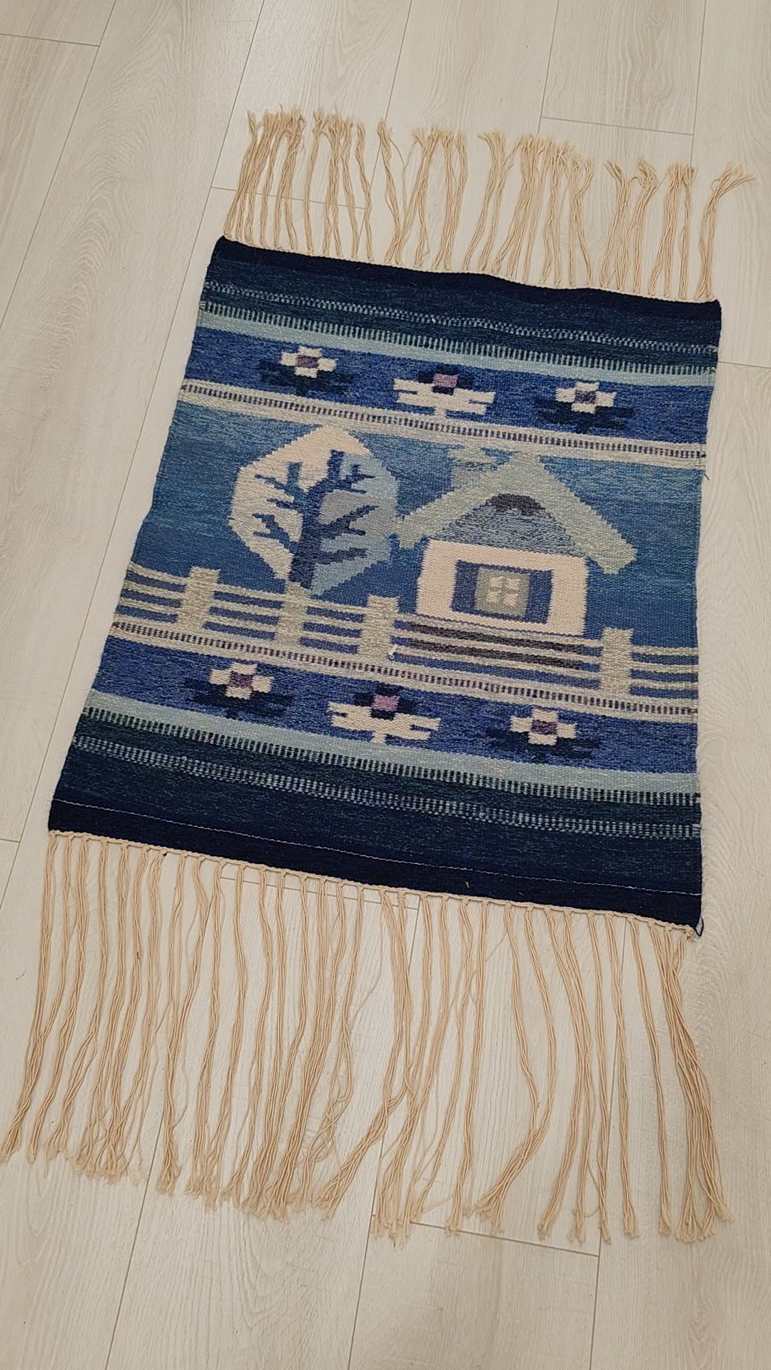 Video showcasing the vintage handwoven wool tapestry laid flat. Closing up on the detailed geometric design of the small house and the tree motives while demonstrating the soft wool texture of the high quality flatweave.