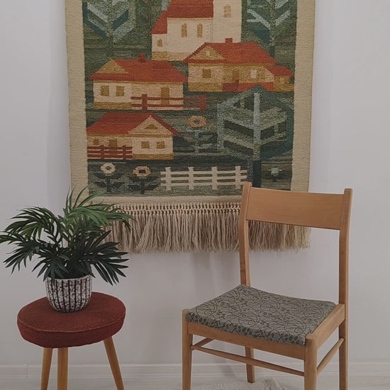 A detailed, sweeping video highlighting a gorgeous vintage 1970s handwoven tapestry with a geometric green and red village scene, showing hands gently ruffling the long macrame fringes.