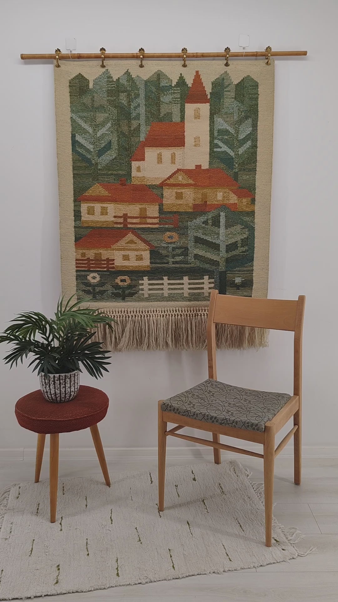 A detailed, sweeping video highlighting a gorgeous vintage 1970s handwoven tapestry with a geometric green and red village scene, showing hands gently ruffling the long macrame fringes.