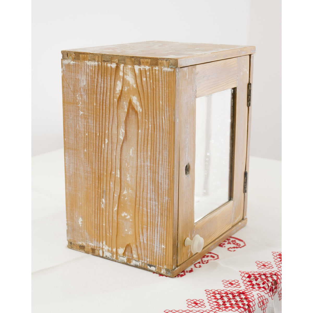 A rustic wooden cabinet with a carefully refinished, distressed wooden structure and a mirror door, shown on a white and red embroidered linen tablecloth - perfect french cottage vibe.