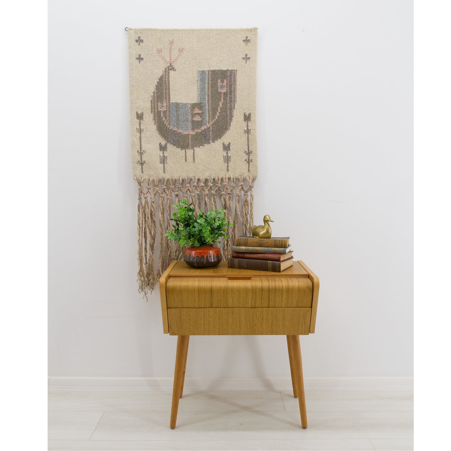 Vintage handwoven wool Scandinavian tapestry displayed on a white wall above a mid-century modern oak side table and plant. Neutral boho home decor.