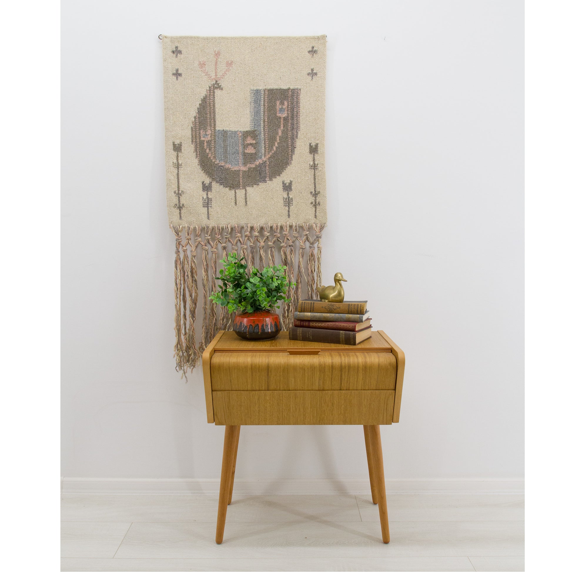 Vintage handwoven wool Scandinavian tapestry displayed on a white wall above a mid-century modern oak side table and plant. Neutral boho home decor.