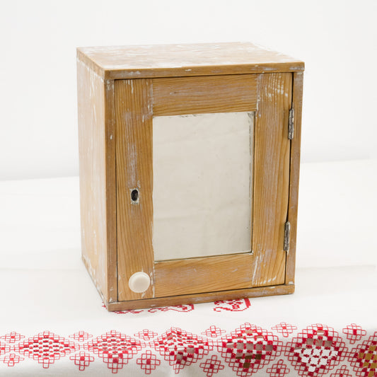 Small rustic medicine cabinet with mirror door, in a beautifully restored vintage condition. Smooth, time-worn finish, a small white knob, and original hinges on the door, set against a white background with a red and white embroidered textile at the bottom.