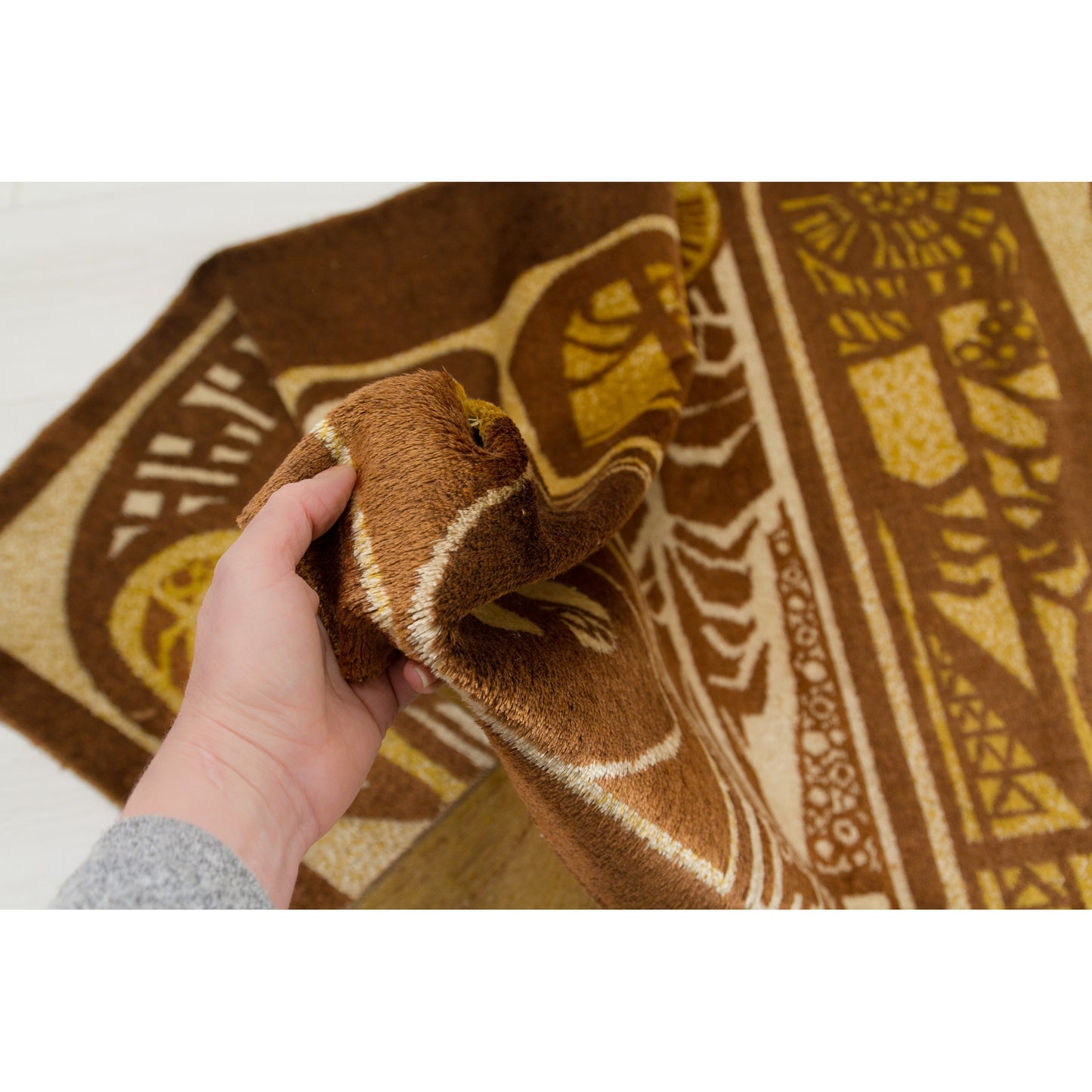 Hand holding the tapestry fabric to demonstrate the extreme softness and flexibility of the vintage moquette pile. Rich, velvety texture in warm earth tones.