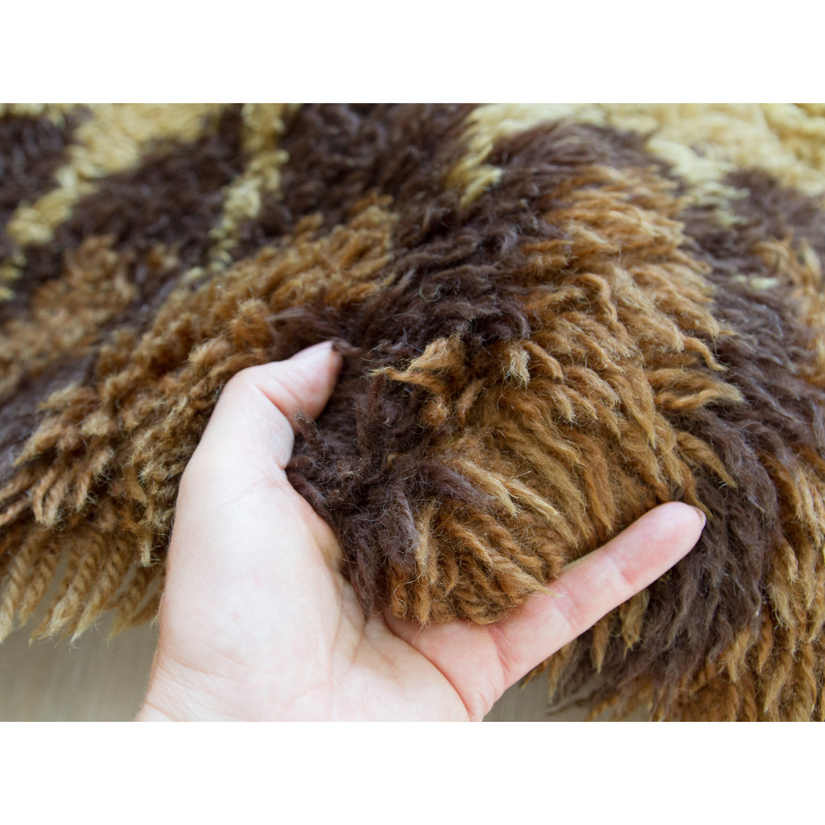 A close-up of a hand holding a chunky, shaggy piles of a vintage rya rug with alternating stripes of shades of brown - a typical 60s / 70s scandinavian color palette.