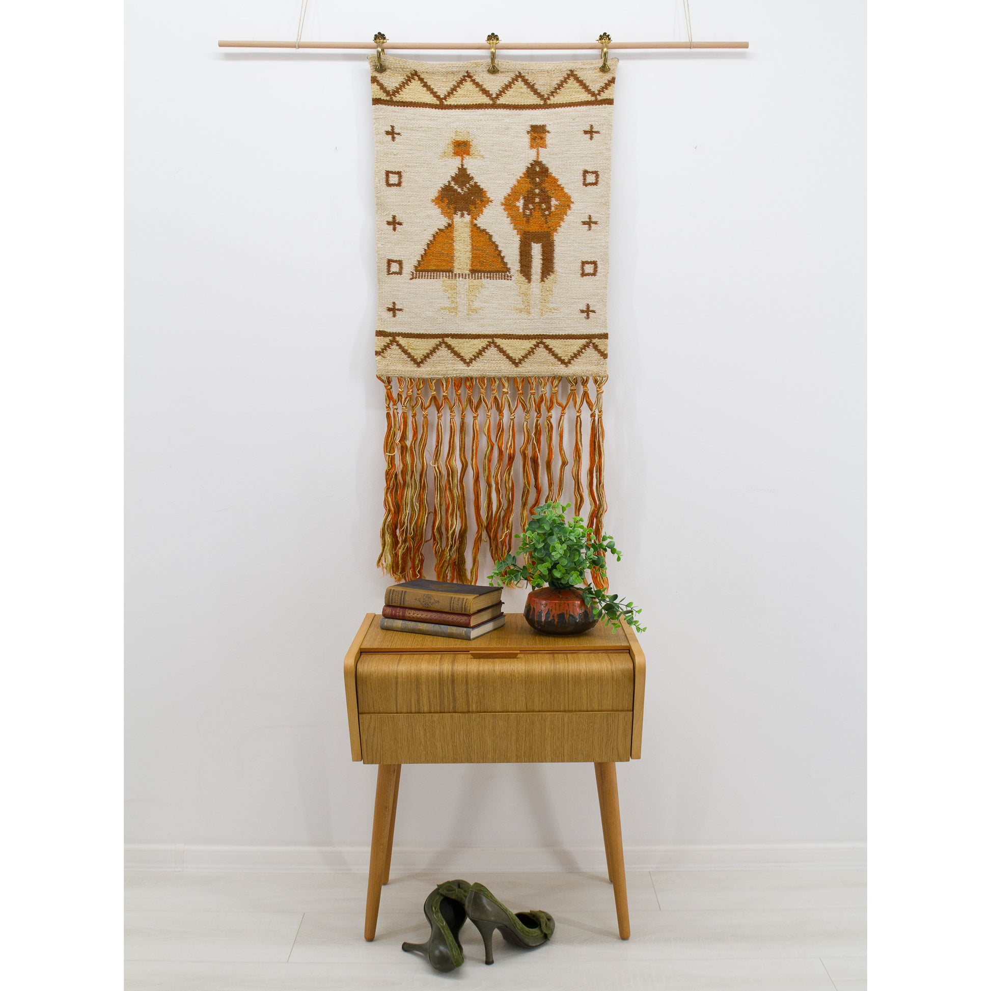 A vintage 1970s warm brown and orange woven wall hanging with long macrame fringes styled beautifully over a white mid-century side table.