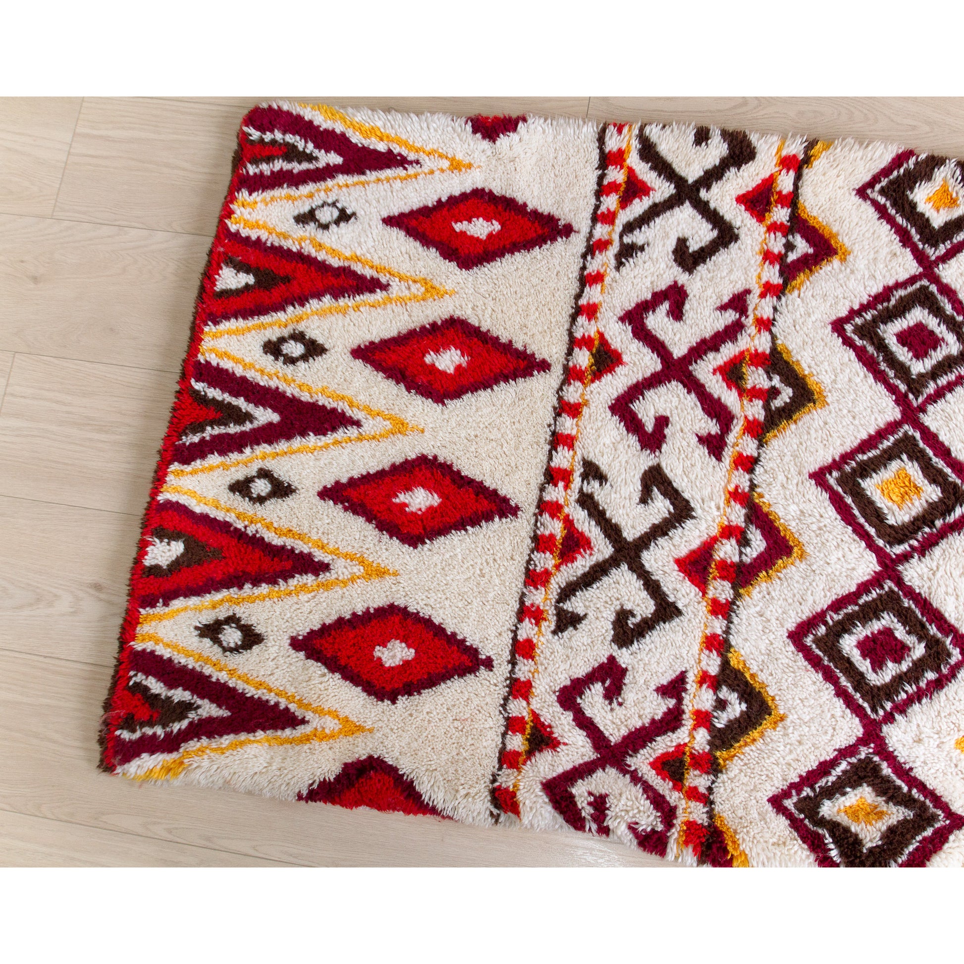 Top down view of the left half of a vintage handmade latch hook rug. Showcasing deep reds, bright burgundy, and gold tribal geometric diamond patterns on pristine white.