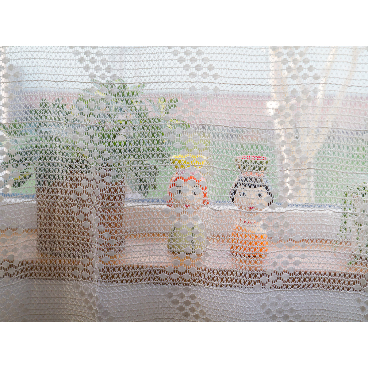 Soft daylight through vintage lace curtain with wooden figurines and plants on the windowsill behind