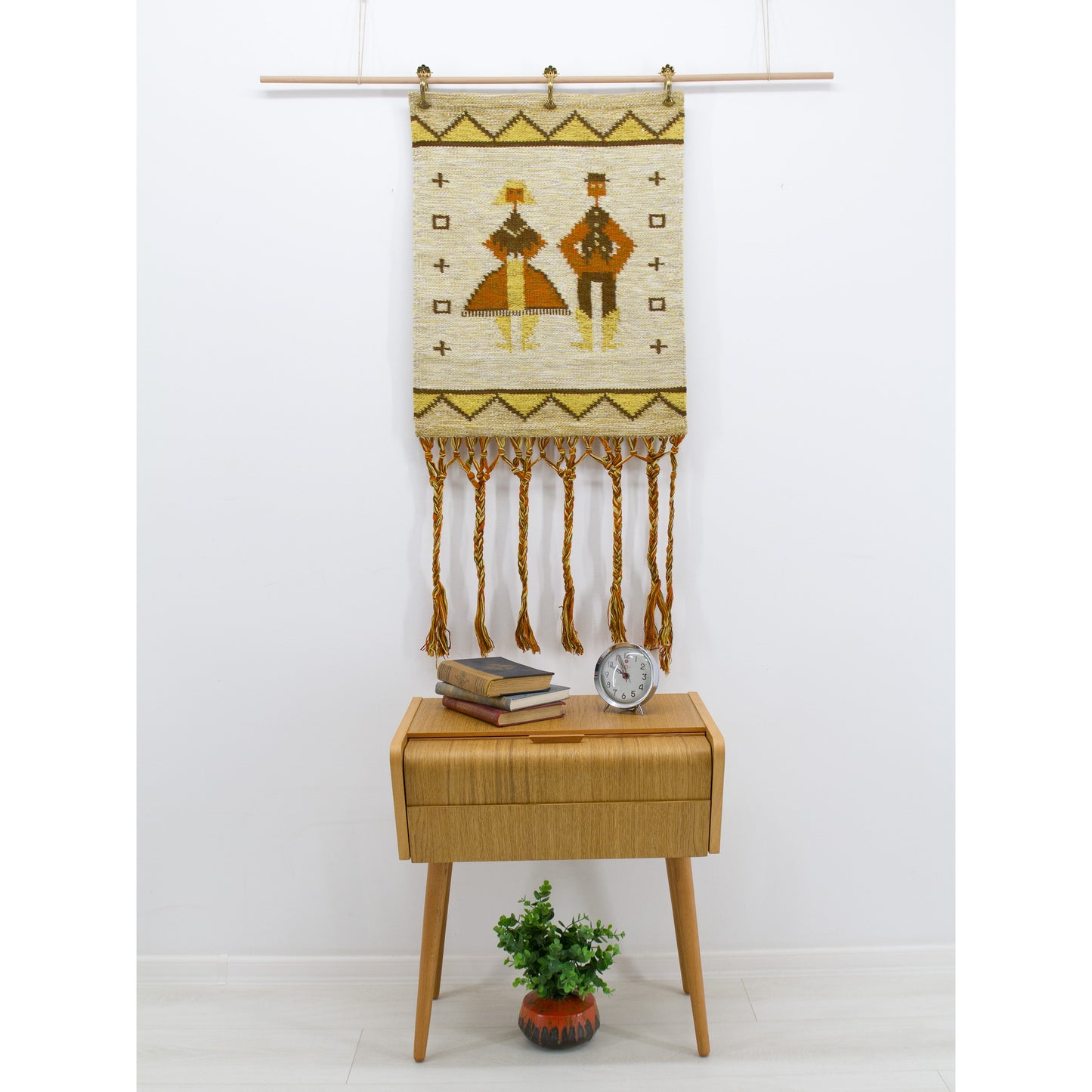A vintage 1970s Eva Nemeth woven tapestry with braided fringes hanging elegantly on a white wall above a retro mid-century teak side table.