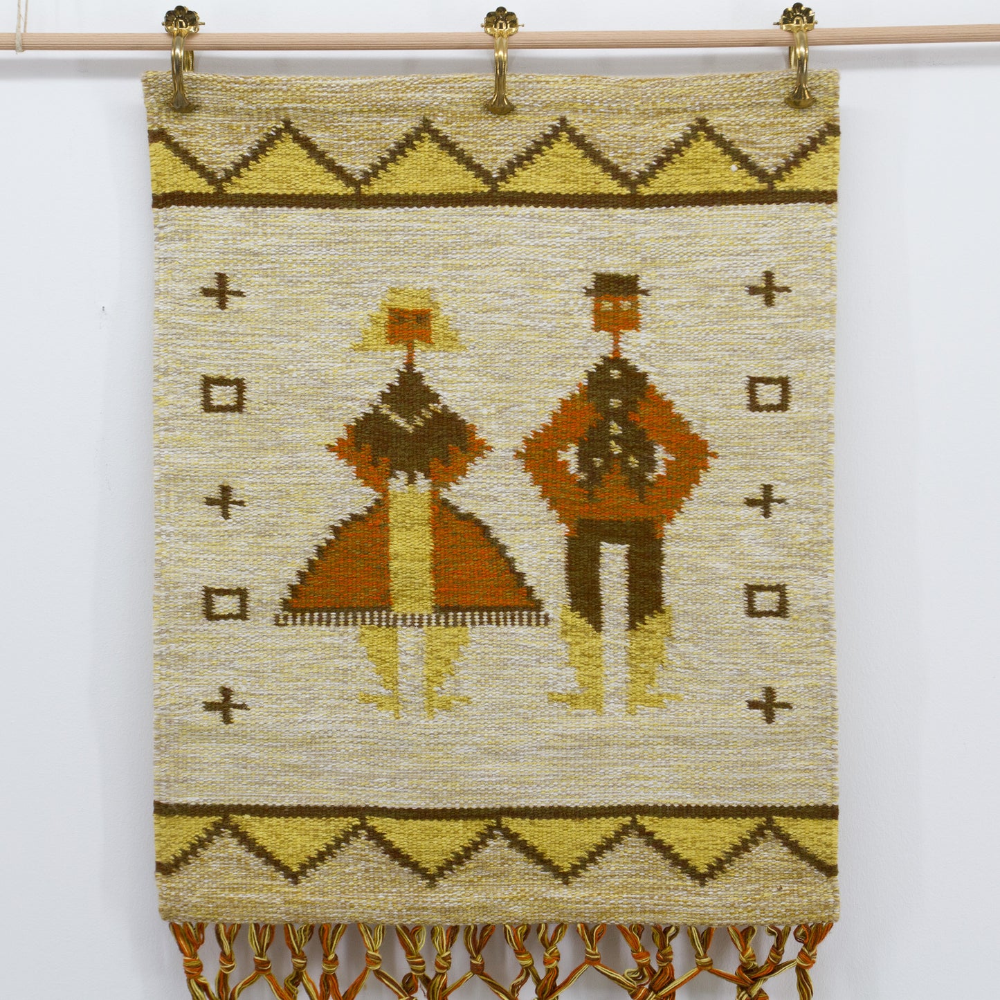 Straight-on, well-lit view focusing entirely on the beautifully woven flatweave section of the vintage folk tapestry, showing stylized geometric human figures in mustard yellow and terracotta, styled gracefully using brass hanging clips.